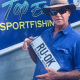 Metanoia Rays Participant Richard Crookes wearing a broad hat and dark sunglasses standing in front of his boat containing the text 'Top End Sport Fishing', holding a Northern Territory number plate containing the phrase "RU. OK".