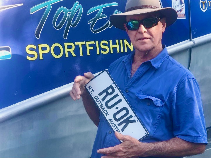 Metanoia Rays Participant Richard Crookes wearing a broad hat and dark sunglasses standing in front of his boat containing the text 'Top End Sport Fishing', holding a Northern Territory number plate containing the phrase "RU. OK".
