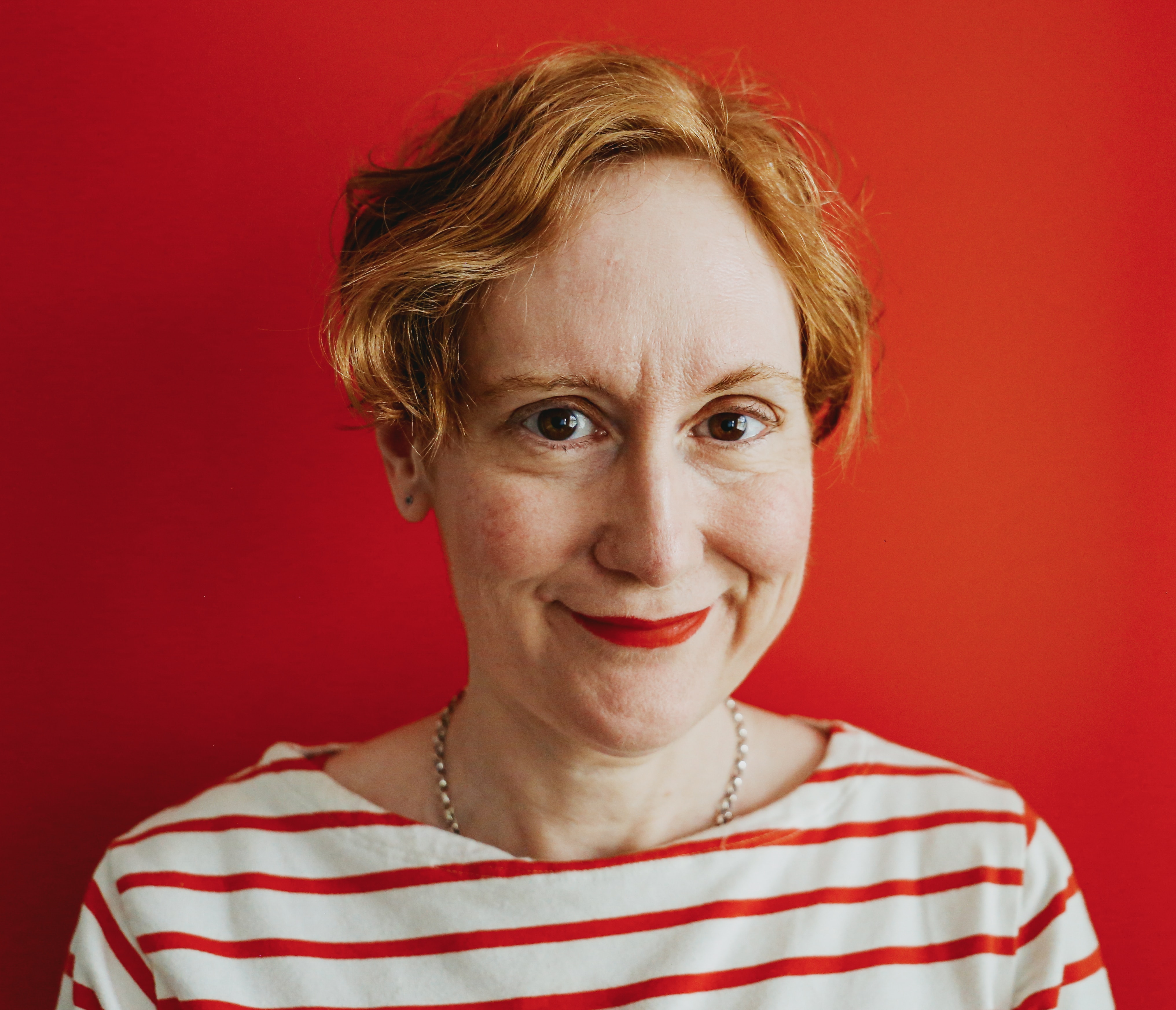 Headshot of female with short orange coloured hair wearing a white and red striped top in front of red background.