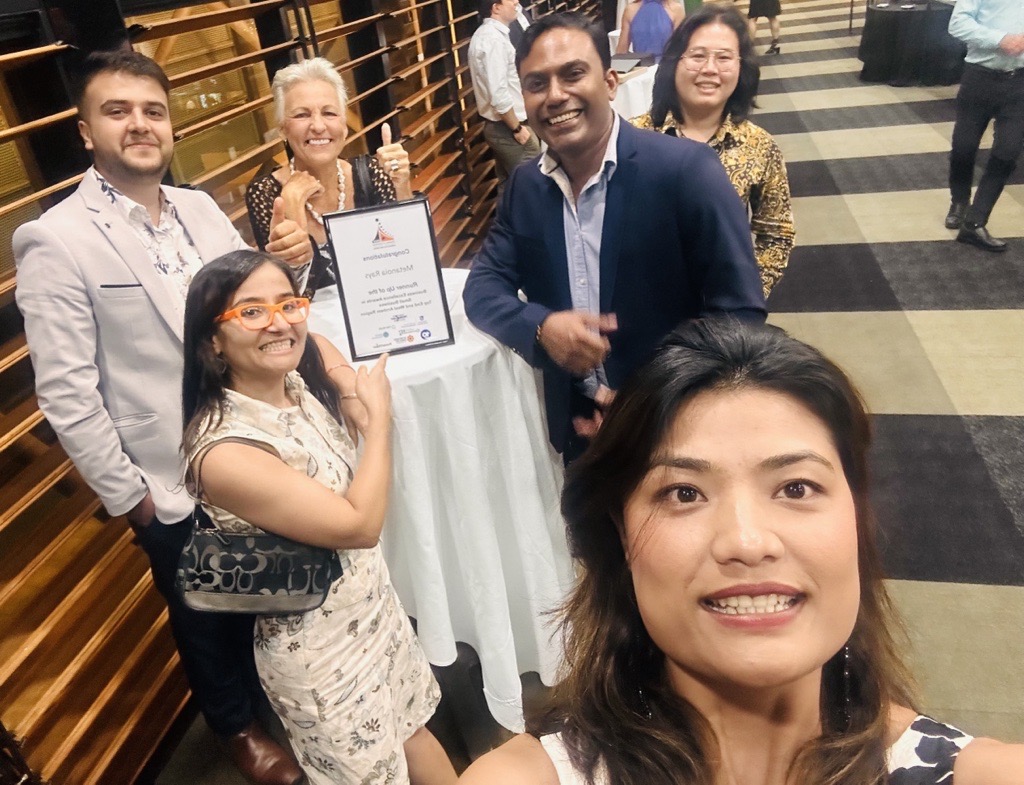 Six members of the Metanoia Rays Team standing around the table with the framed certificate on the table at the 2023 Northern Territory Chamber of Commerce Business Excellence Awards.